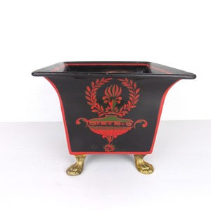 Peut inclure: Un cache-pot carré noir avec un bord évasé et une bordure peinte en rouge. L'extérieur présente un motif d'urne décoratif rouge et vert avec des feuilles de laurier. Le cache-pot repose sur quatre pieds en métal doré en forme de pattes.