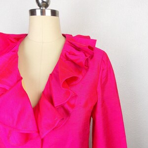 Pu&ograve; includere: Un blazer rosa shocking con un grande colletto a balze. Il blazer ha maniche lunghe e una chiusura con un solo bottone.