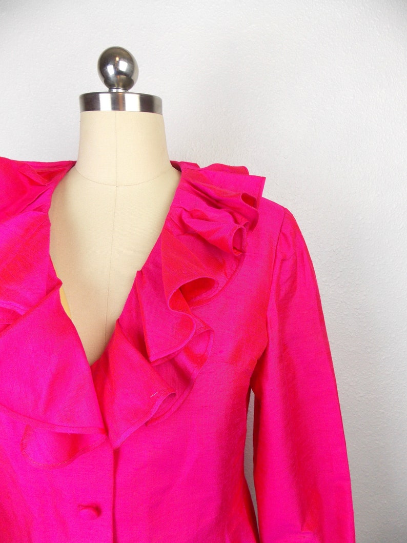 Pu&ograve; includere: Un blazer rosa shocking con un grande colletto a balze. Il blazer ha maniche lunghe e una chiusura con un solo bottone.