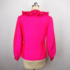 Pu&ograve; includere: Una blusa rosa brillante con un colletto a volant e maniche lunghe. La blusa &egrave; su uno sfondo bianco.