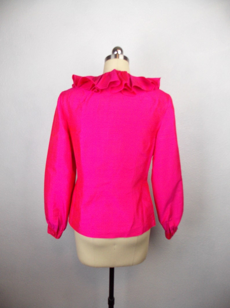 Pu&ograve; includere: Una blusa rosa brillante con un colletto a volant e maniche lunghe. La blusa &egrave; su uno sfondo bianco.