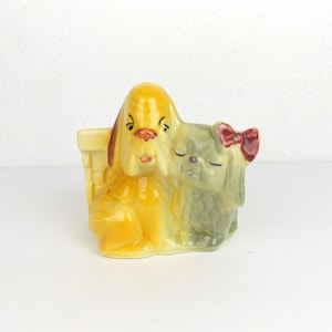 May include: A ceramic planter featuring two dog figurines. One dog is yellow with brown ears, and the other is green with a red bow. The planter has a basket-like design. The dogs are side-by-side, creating a charming decorative piece.