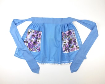 May include: A blue apron with white trim and two floral print pockets. The apron has long ties that can be tied around the waist.