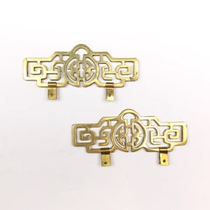 May include: Two matching brass furniture pulls featuring an intricate openwork geometric pattern with a central circular motif and two downward-facing mounting tabs on the bottom edge of each piece, showcasing a polished metallic finish.