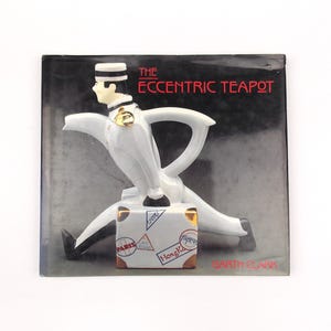 May include: A white ceramic teapot in the shape of a bellhop with a suitcase. The teapot has a black and white hat, a gold emblem, and the words "THE ECCENTRIC TEAPOT" in red. The suitcase has travel stickers.