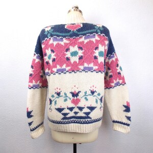 May include: Cream-colored sweater with a colorful geometric pattern. The sweater features a navy blue neckline and accents, with pink, purple, and teal floral and diamond designs. The sleeves and lower portion of the sweater are cream-colored.
