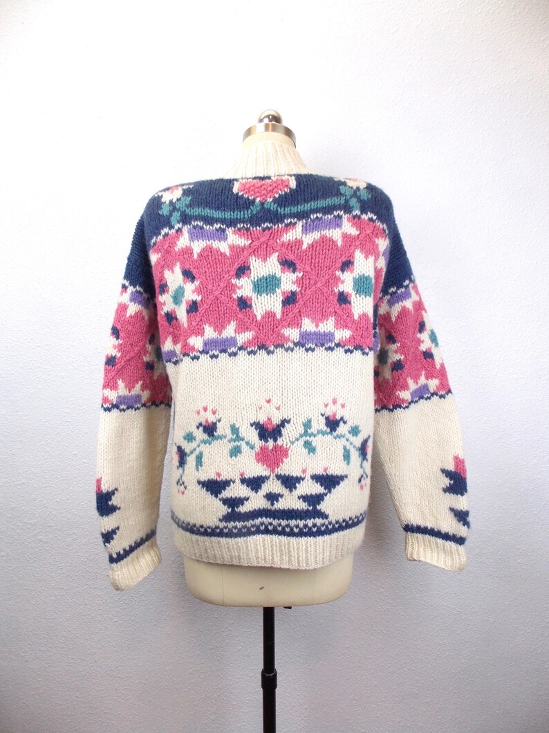 May include: Cream-colored sweater with a colorful geometric pattern. The sweater features a navy blue neckline and accents, with pink, purple, and teal floral and diamond designs. The sleeves and lower portion of the sweater are cream-colored.