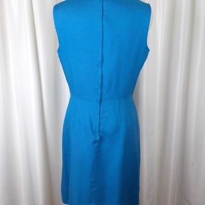 May include: A sleeveless, knee-length turquoise dress displayed on a mannequin. The dress features a fitted bodice, a slight A-line skirt, and a vertical seam down the back. The fabric has a subtle texture, and the dress is set against a white curtain backdrop.