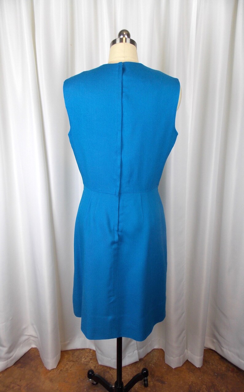 May include: A sleeveless, knee-length turquoise dress displayed on a mannequin. The dress features a fitted bodice, a slight A-line skirt, and a vertical seam down the back. The fabric has a subtle texture, and the dress is set against a white curtain backdrop.