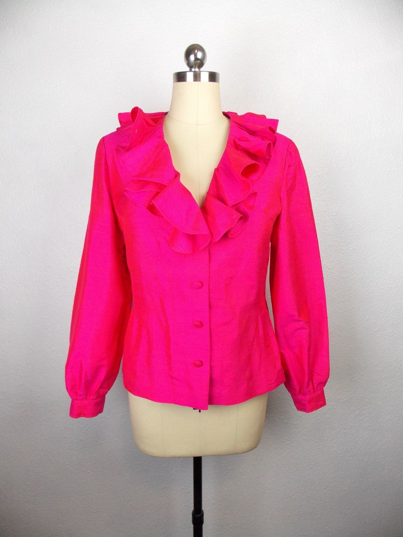 Pu&ograve; includere: Una blusa rosa brillante con un grande colletto a volant e maniche lunghe. La blusa ha una chiusura con bottoni sul davanti.
