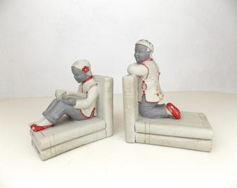 May include: Pair of vintage bookends featuring figures in traditional attire. One figure is seated, reading a book, while the other is kneeling. Both figures are dressed in white jackets with red trim, gray pants, and red shoes. The bookends are gray and shaped like stacked books.