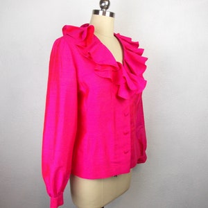 Pu&ograve; includere: Una blusa rosa shocking con un grande colletto a balze e maniche lunghe. La blusa ha una chiusura con bottoni sul davanti.