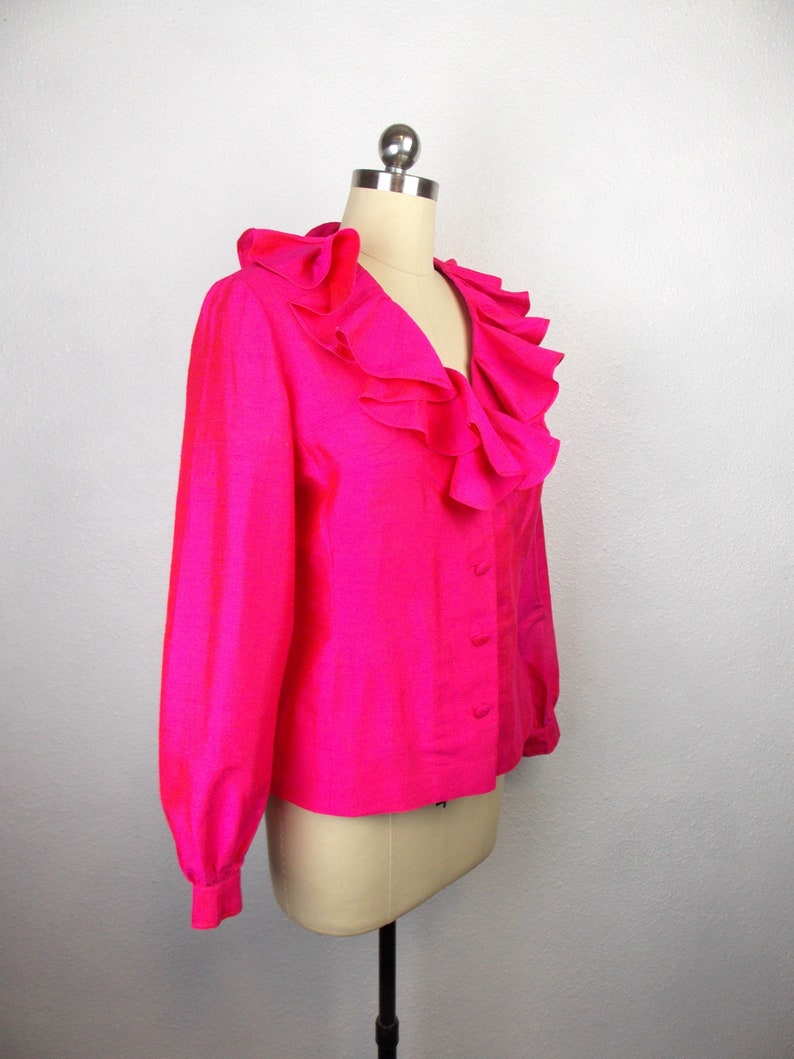 Pu&ograve; includere: Una blusa rosa shocking con un grande colletto a balze e maniche lunghe. La blusa ha una chiusura con bottoni sul davanti.