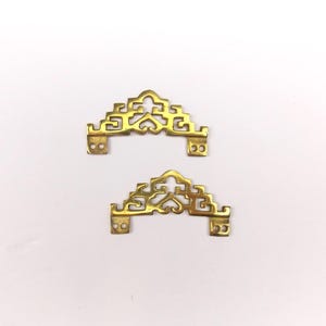 May include: Two matching brass furniture corner plates featuring an intricate geometric cutout pattern. Each piece has a curved top edge and two small circular screw holes on each side for mounting to a flat surface.