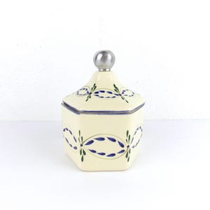 May include: Cream-colored ceramic jar with a silver knob on the lid. The jar has a hexagonal shape and is decorated with blue and green floral designs. The lid is outlined in blue.