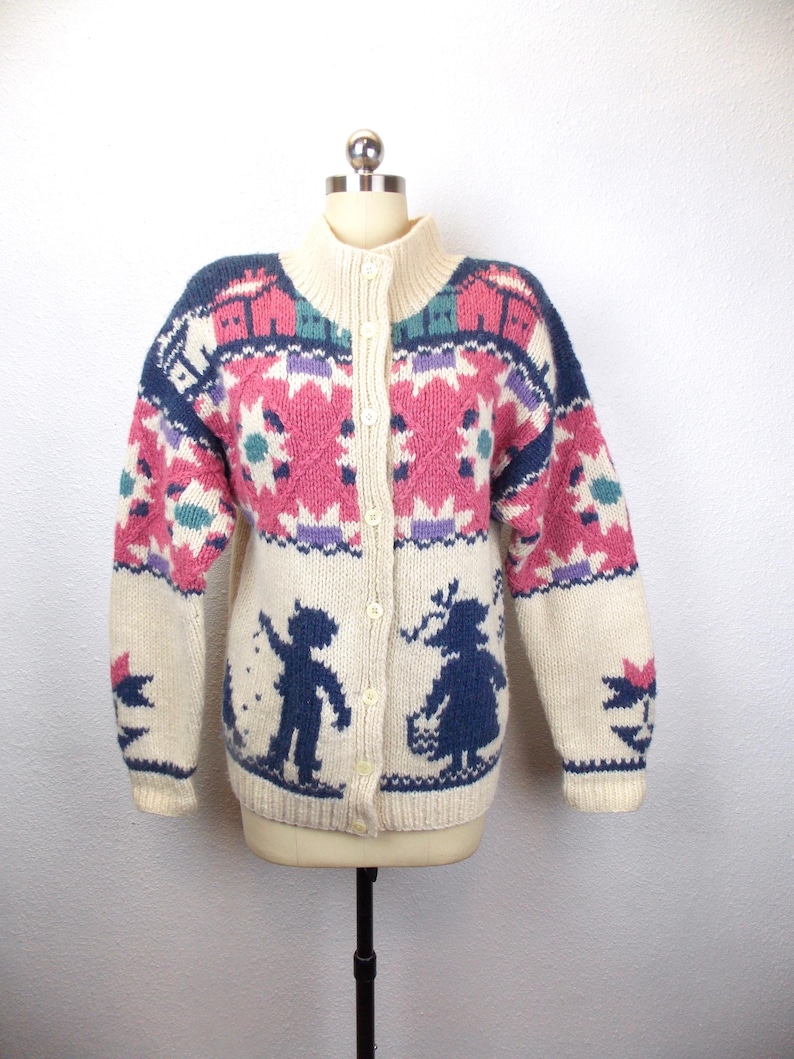 May include: Cream-colored, button-down cardigan sweater with a high collar. The sweater features a colorful geometric pattern in pink, blue, and purple. Silhouettes of a boy and girl are on the front. The sweater is displayed on a mannequin.