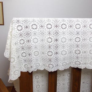 May include: An off-white lace tablecloth featuring a repeating square pattern of circular floral motifs. The fabric has a delicate, open-work design with a scalloped edge along the bottom border, showcasing intricate needlework and fine thread detail.