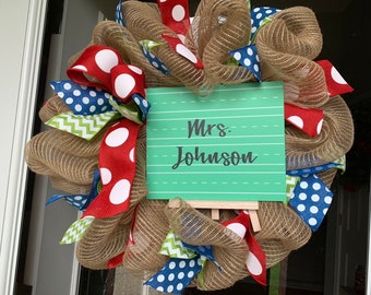 May include: A burlap wreath decorated with red, blue, and green polka dot ribbon. A green chalkboard sign with the text "Mrs. Johnson" is attached to the wreath.