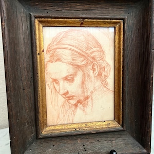 May include: A framed sketch of a woman's face, rendered in reddish-brown tones. The artwork is set within a gold-lined frame, encased by a dark, rustic wooden frame. The piece is displayed on a black easel.