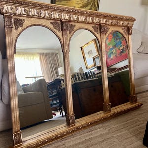 May include: A large, rectangular, gold-toned mirror featuring three arched glass panels separated by ornate, fluted columns. The frame is decorated with intricate carvings, including floral motifs at the top of each arch and a detailed base.
