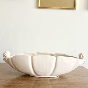 May include: Cream-colored ceramic decorative bowl with a scalloped edge and two scrolled handles. The bowl has a ribbed design and sits on a wooden surface. Decorative home decor.