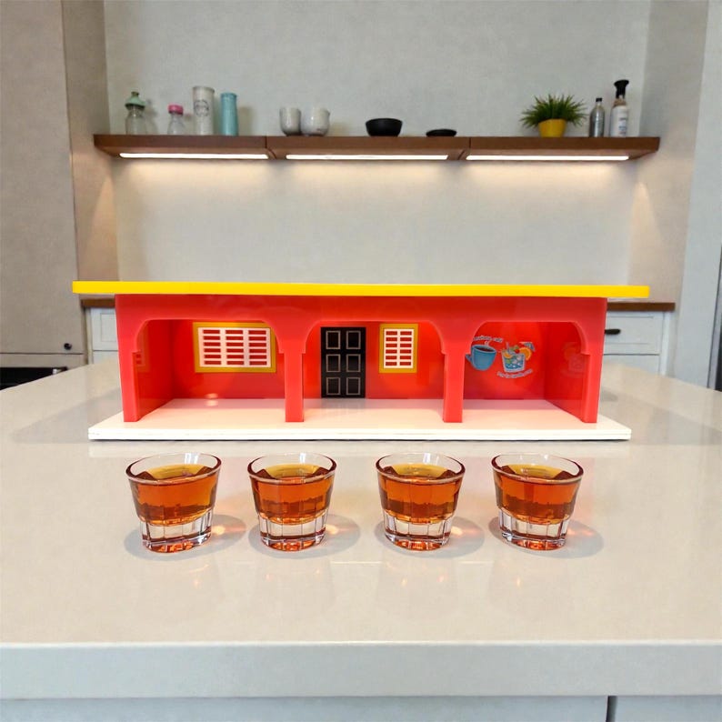 May include: A red and yellow toy building with four shot glasses filled with amber liquid. The building has a yellow roof, red walls, and a white base. The shot glasses are clear and sit on a white surface.