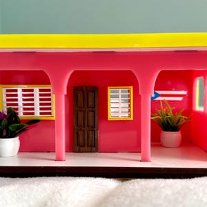 May include: A miniature pink house with a yellow roof, arched supports, and a brown door. The model features two windows with white shutters, potted plants, and a Puerto Rican flag. The base is brown and the house is on a white surface.
