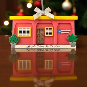 May include: A pink and yellow miniature house ornament with a Puerto Rican flag and the text "No Me Quiero Ir De Aquí". The ornament has a silver ribbon for hanging and is decorated with small green plants.