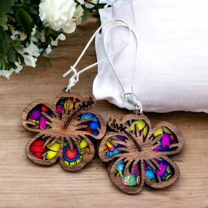 Puede incluir: Un par de pendientes de madera con forma de flores de hibisco. Los pendientes están pintados con un diseño abstracto colorido y tienen ganchos plateados.