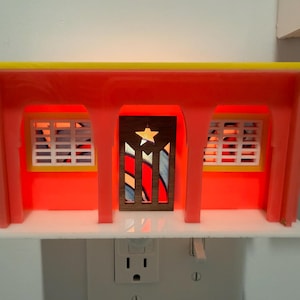May include: A decorative night light shaped like a building, with a red and yellow design. The centre door has a star and flag design. The windows have a white grid pattern. The light is plugged into a wall socket.