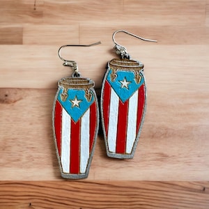 May include: A pair of silver earrings shaped like Puerto Rican congas, painted with the Puerto Rican flag. The congas are red, white, and blue with a gold star on the blue stripe.