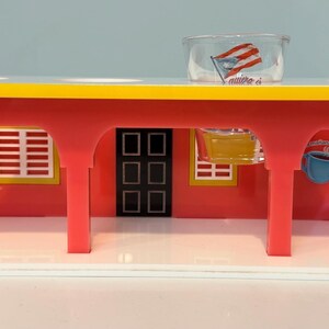 May include: A red and yellow toy diner with a white base. The diner has a yellow roof, red walls, and a black door. A clear plastic cup with a flag is on top. The diner has two windows and a mural of a drink.
