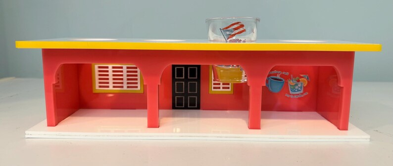 May include: A red and yellow toy diner with a white base. The diner has a yellow roof, red walls, and a black door. A clear plastic cup with a flag is on top. The diner has two windows and a mural of a drink.