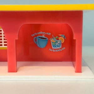 May include: A red and yellow toy structure with a white base. The red interior features illustrations of a coffee cup and a cocktail with text that reads "Por la ma&ntilde;ana, caf&eacute;" and "Por la tarde, ron."