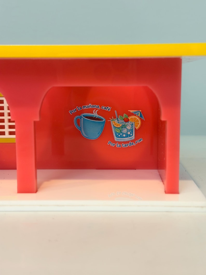 May include: A red and yellow toy structure with a white base. The red interior features illustrations of a coffee cup and a cocktail with text that reads "Por la ma&ntilde;ana, caf&eacute;" and "Por la tarde, ron."