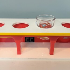 May include: A colorful, multi-holed serving tray with a white top, red supports, and a yellow edge. A small glass bowl sits in one of the holes. The tray is designed to hold multiple items.