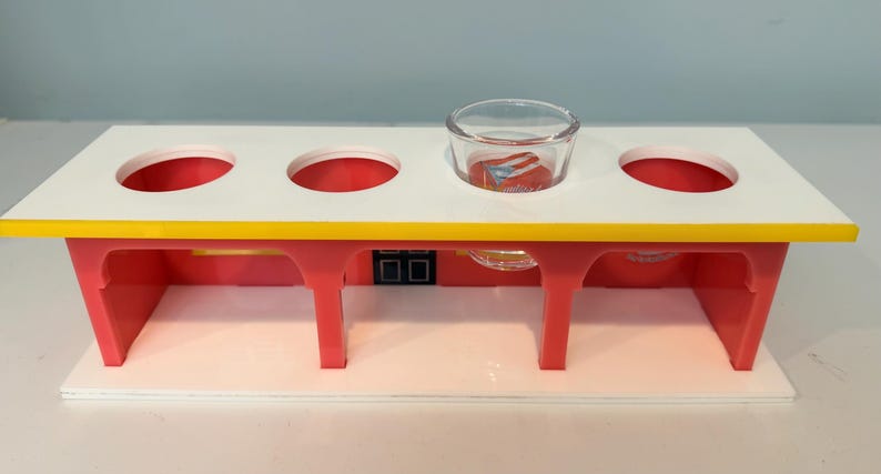 May include: A colorful, multi-holed serving tray with a white top, red supports, and a yellow edge. A small glass bowl sits in one of the holes. The tray is designed to hold multiple items.