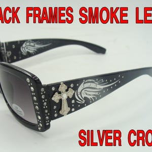 May include: Black sunglasses with smoke lenses. The frames are adorned with rhinestones and a silver cross. The arms feature a decorative wing design. Text on the image reads "BLACK FRAMES SMOKE LENS" and "SILVER CROSS".
