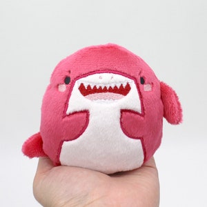 May include: A pink plush toy shaped like a shark with white embroidered eyes, teeth, and a belly. The shark has a happy expression.