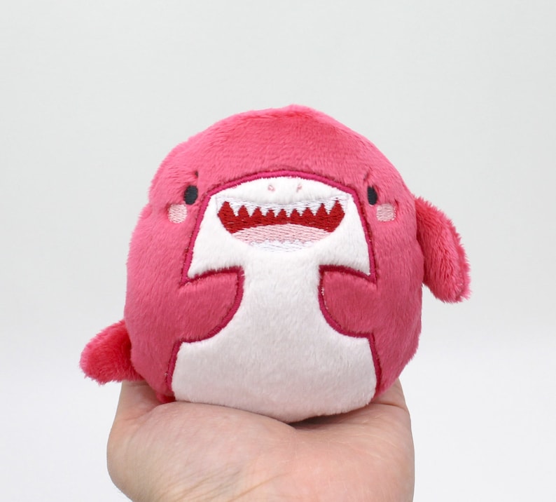 May include: A pink plush toy shaped like a shark with white embroidered eyes, teeth, and a belly. The shark has a happy expression.