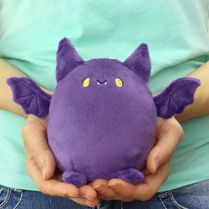 Può includere: Un pipistrello di peluche viola con occhi gialli e un piccolo sorriso. Il pipistrello ha grandi ali e piccoli piedi.