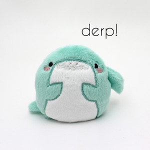 May include: A small, plush, light green and white toy with embroidered eyes and a mouth. The toy is shaped like a round, chubby animal with a small fin on the side. The text "derp!" is written above the toy.