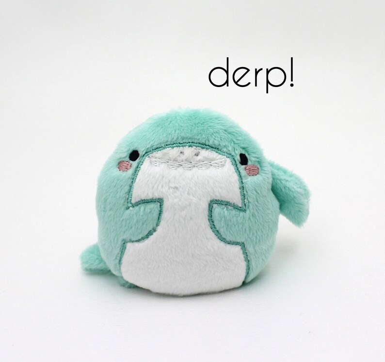 May include: A small, plush, light green and white toy with embroidered eyes and a mouth. The toy is shaped like a round, chubby animal with a small fin on the side. The text "derp!" is written above the toy.