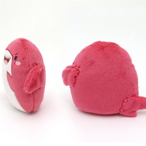 May include: A plush pink shark toy with a white belly and a cute embroidered face. The shark has a fin on each side and a tail.