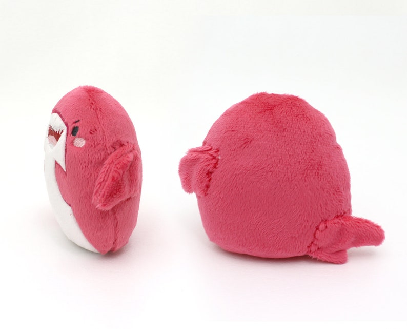 May include: A plush pink shark toy with a white belly and a cute embroidered face. The shark has a fin on each side and a tail.