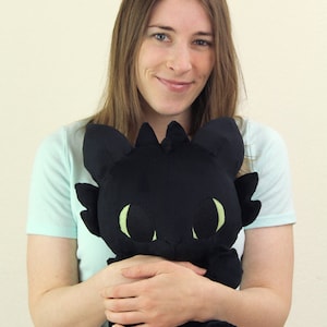 May include: A woman is holding a black plush toy dragon with green eyes. The dragon is about the size of a small child. The woman is smiling and looking at the camera.