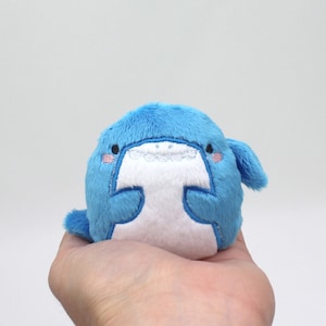 May include: A small, blue plush toy shaped like a whale with white embroidered eyes and a mouth. The whale has a white belly and is holding a small, white heart.