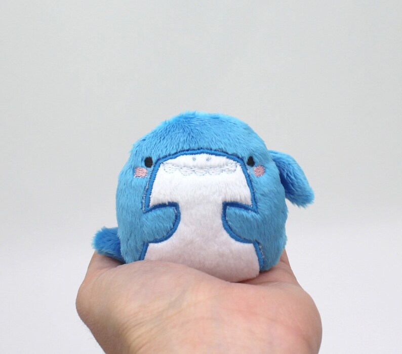 May include: A small, blue plush toy shaped like a whale with white embroidered eyes and a mouth. The whale has a white belly and is holding a small, white heart.