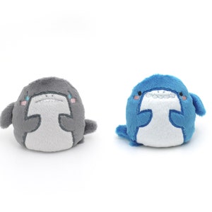 May include: Two plush shark toys, one gray and one blue, with white bellies and embroidered eyes and mouths. The sharks are sitting on a white background.