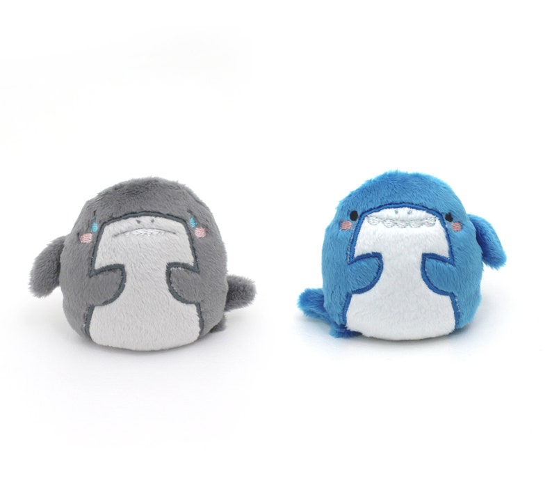 May include: Two plush shark toys, one gray and one blue, with white bellies and embroidered eyes and mouths. The sharks are sitting on a white background.